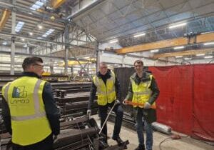 Trabajadores con chalecos de seguridad de pie en un taller industrial rodeados de materiales metálicos.